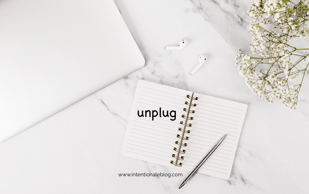 image of a plain white notebook a silver pen and ear buds, notebook with a text "unplug"- as part of intentional productivity 