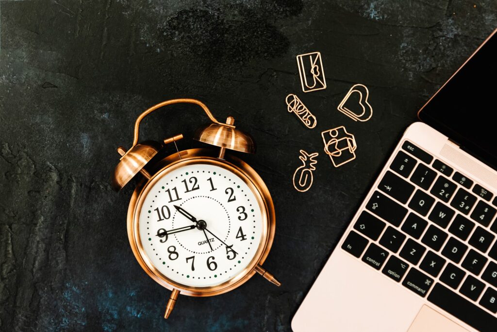 ose-gold-old-fashioned-alarm-clock-with-black-numbering-laid-on-dark-textured-background-with-quirky-shape-rose-gold-paperclips-and-rose-laptop